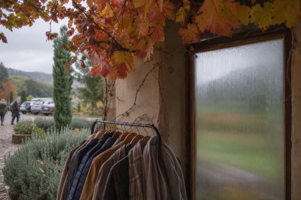 Rack of light jackets and scarves beside a fogged cellar door window with vineyard rows dim in the mist outside
