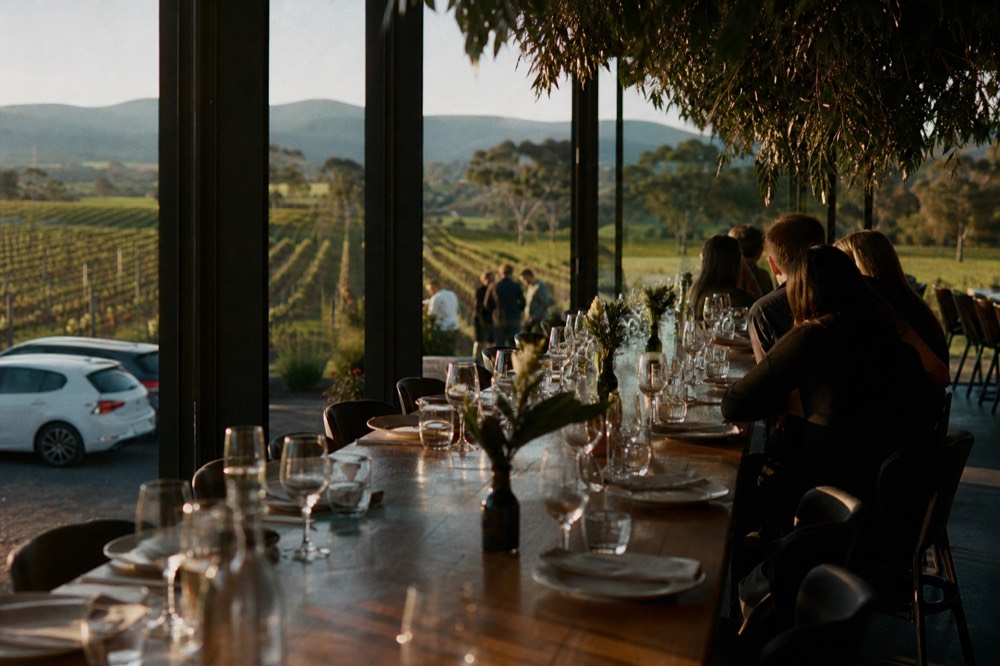 Long communal table set for lunch in a winery restaurant with vineyard visible through tall windows