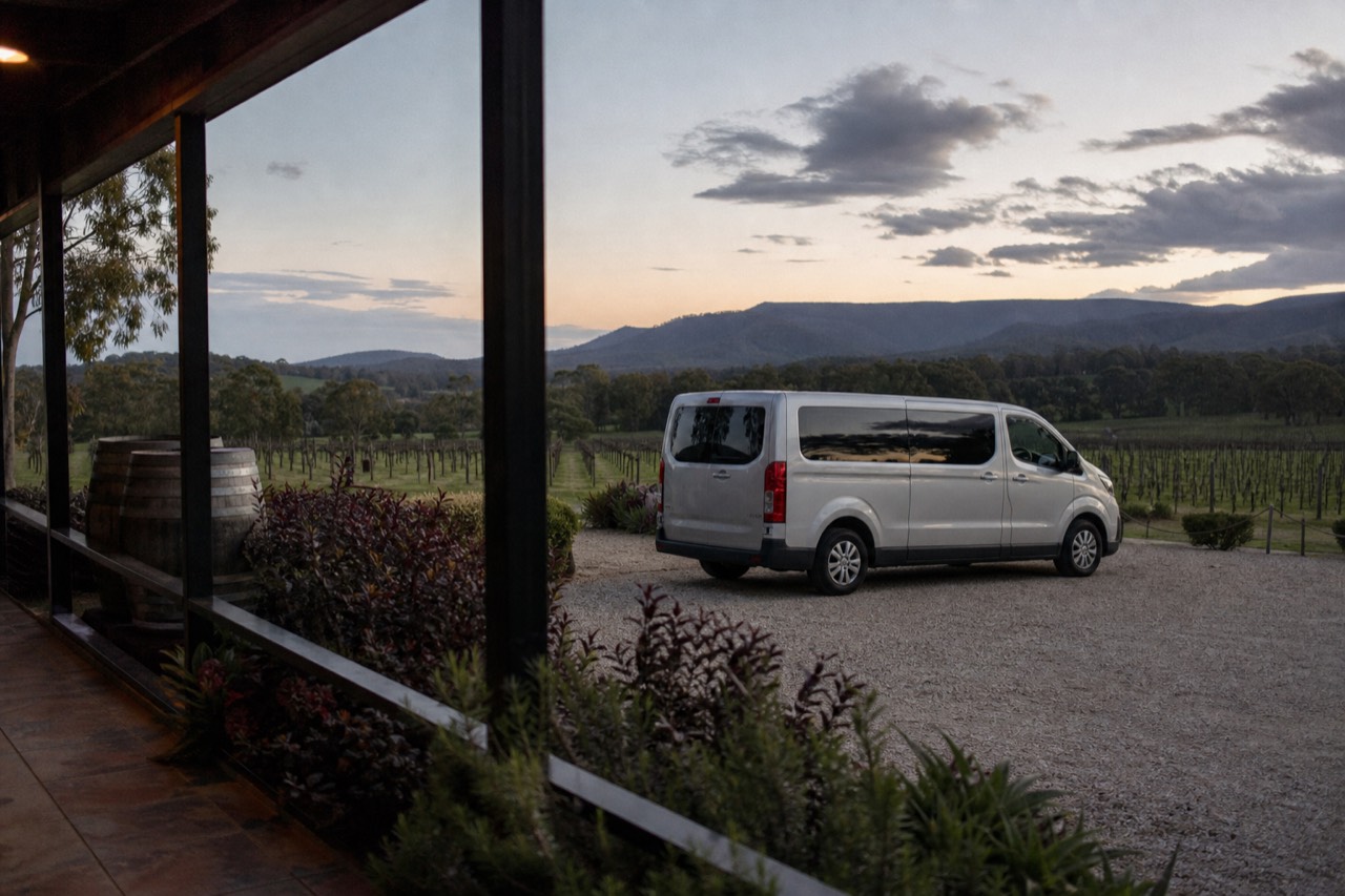 Small tour van parked on gravel beside a cellar door with vineyard rows running away behind it in afternoon light