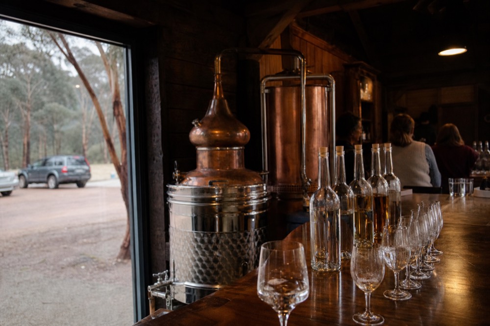 Small copper still and rows of clear bottles in a distillery tasting room with a group of glasses on the counter
