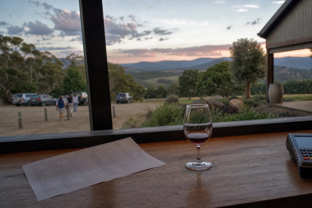 Printed tasting list and a small pour of red wine on a cellar-door counter with a card machine just visible at the edge