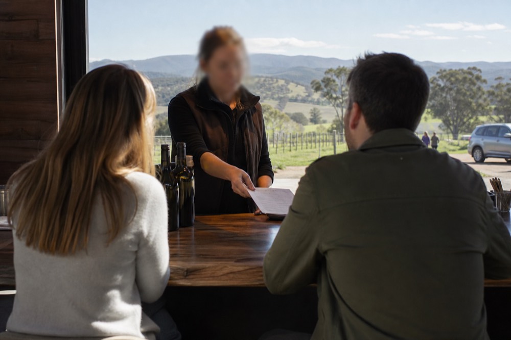 Cellar door host handing a paper tasting list across a timber counter to two seated visitors