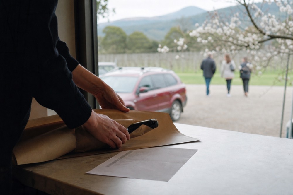 Hands wrapping a single purchased bottle in paper at a cellar door counter beside a printed tasting list