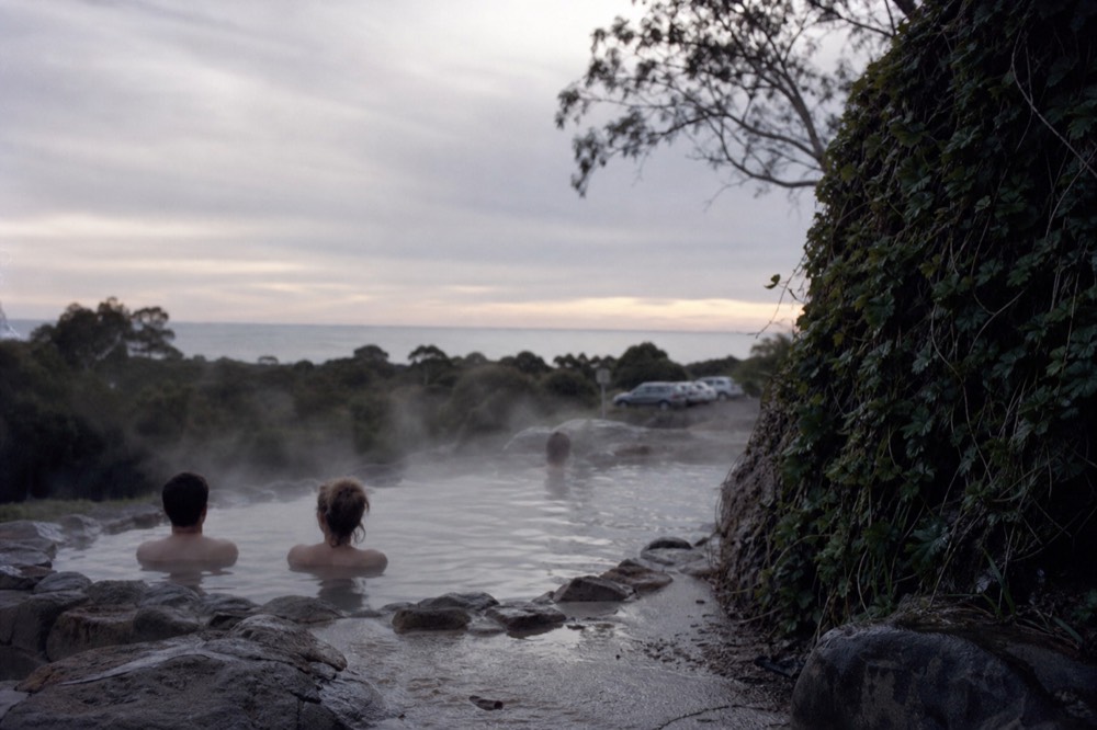 Steam rising from a stone-edged geothermal bathing pool set into a hillside at dusk, bathers seated at the rim, distant coastal treeline