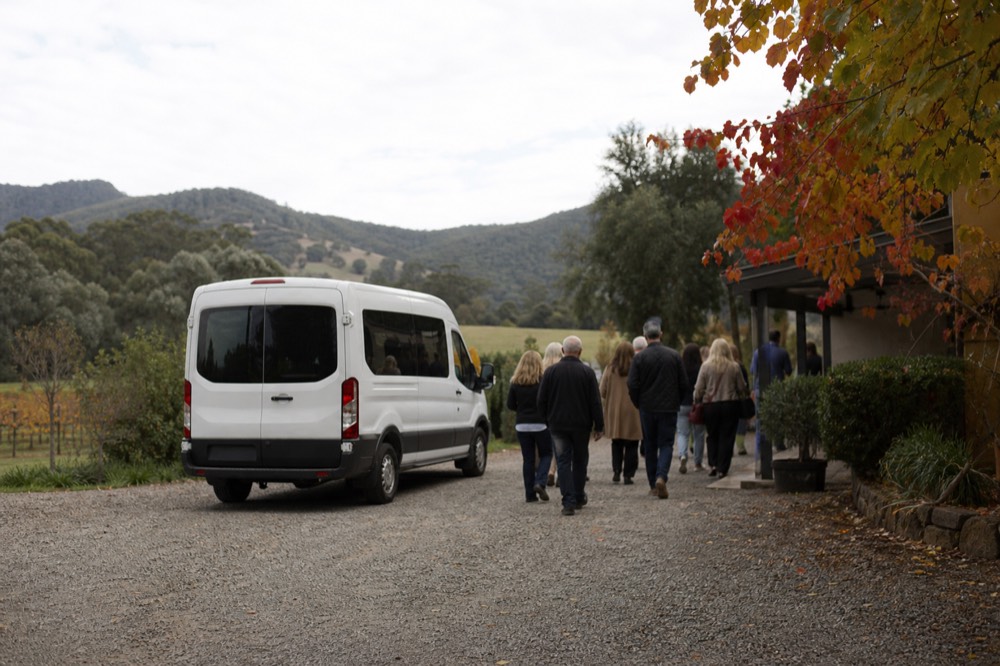 A small tour van parked on gravel outside a cellar door with a group of eight visitors walking toward the entrance