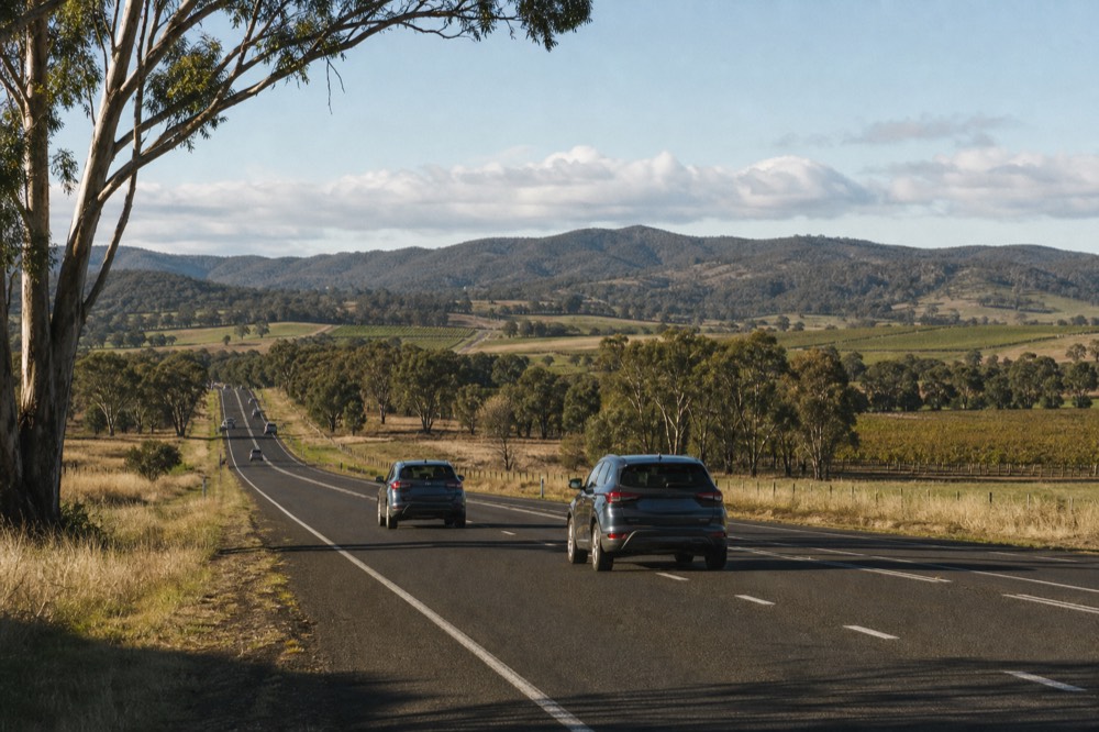 Highway heading out of a city fringe toward green hills, farmland on both sides, mid-morning traffic light
