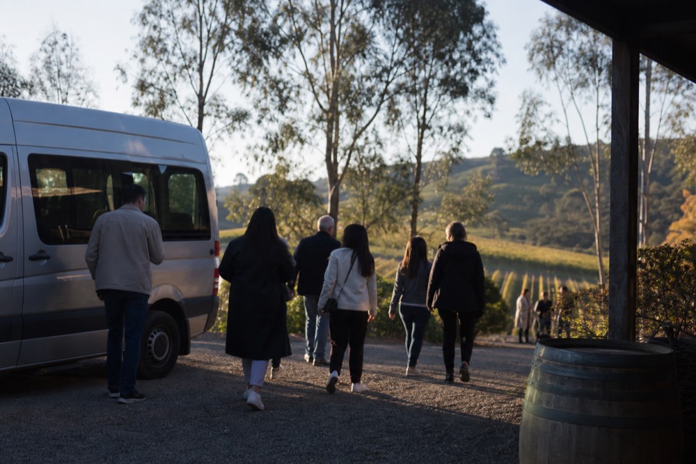 Passengers stepping down from a small tour van onto gravel outside a cellar door in early afternoon light