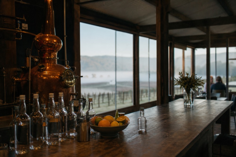 Copper gin still behind a wooden bar with tonic bottles and citrus on the counter in a distillery tasting room
