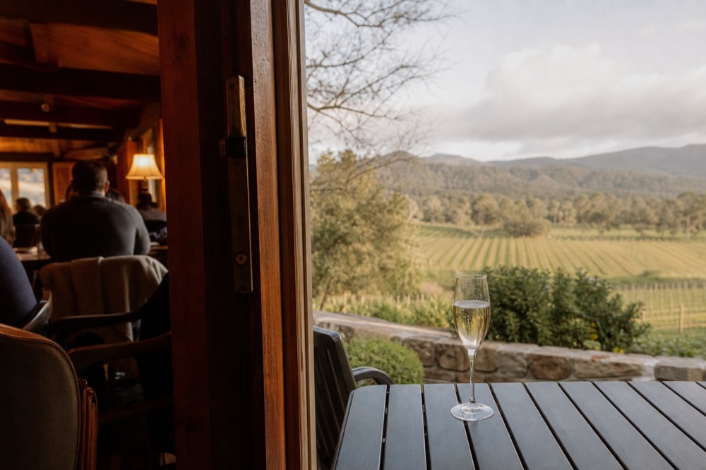 Sparkling wine flute on an outdoor terrace table with vine rows and a distant hillside behind, soft afternoon shadows