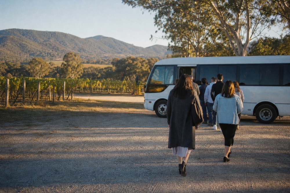 Passengers boarding a small bus at a rural cellar door stop, viewed from behind, gravel car park and vine rows around them