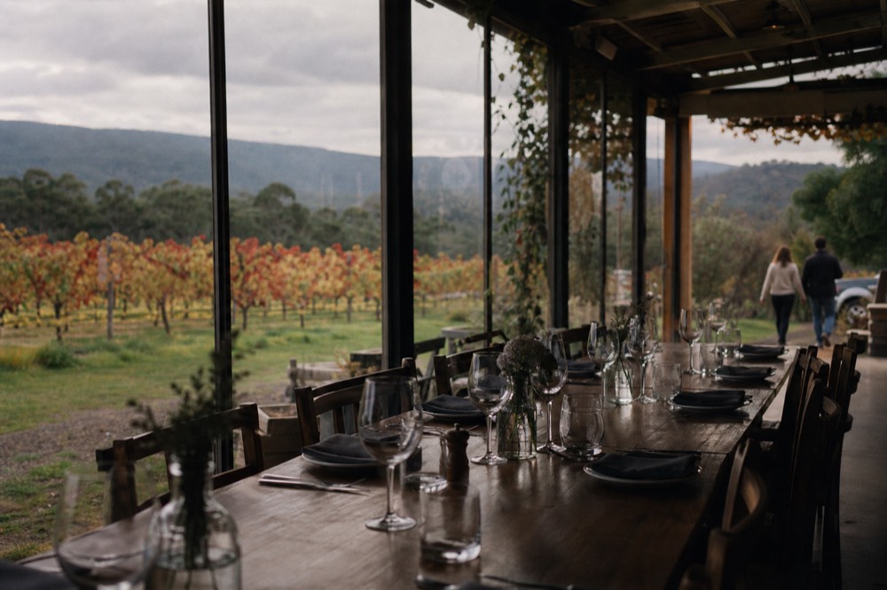 Long communal table set for lunch inside a winery restaurant with windows opening onto vines, plates and wine glasses laid out, no diners seated