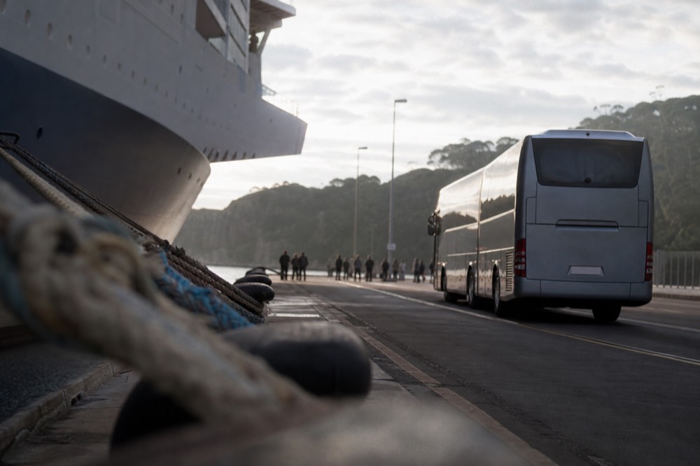 Coach waiting on a wide portside apron at dawn with bollards and mooring lines in the foreground, large ship hull out of focus behind