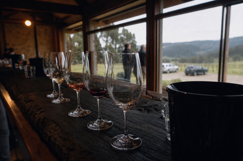 Row of tasting glasses filled to different levels on a slate bench with a spit bucket at the end