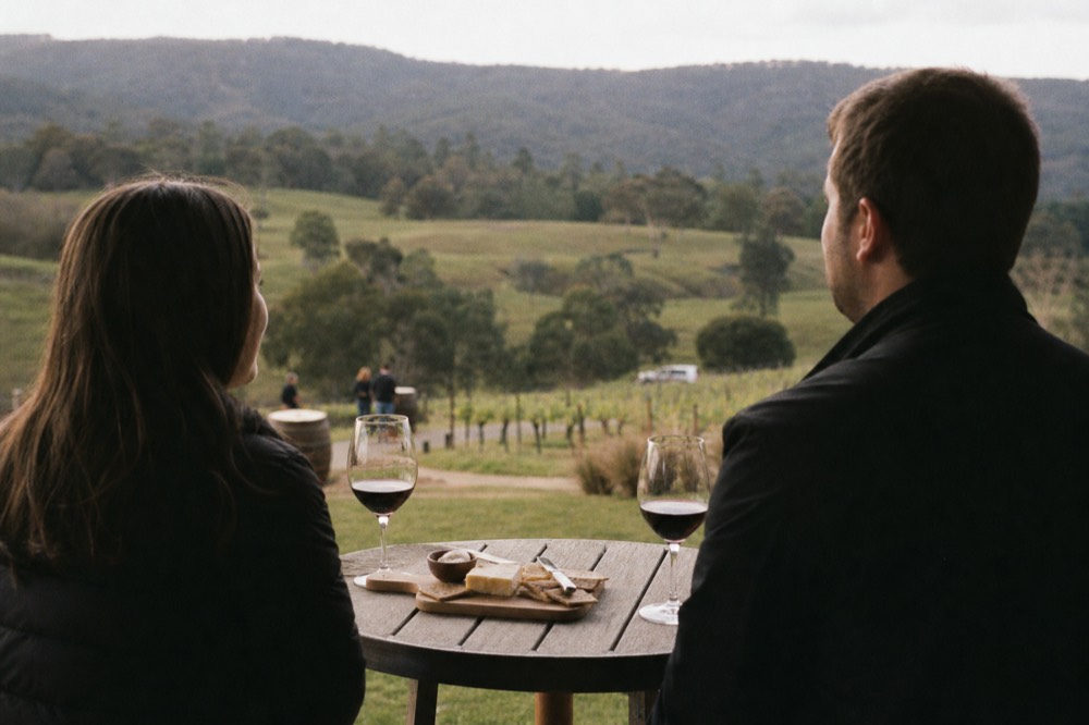 A couple seated at a small outdoor table with two wine glasses and a cheese plate, vineyard slope falling away behind them, late afternoon