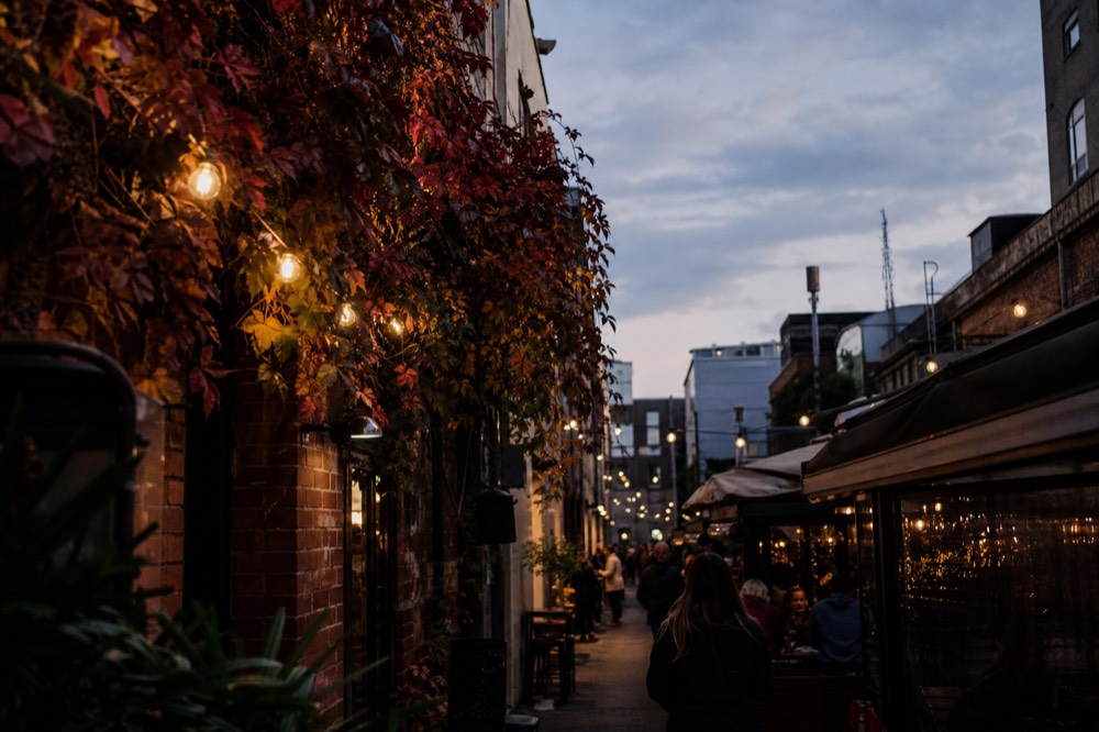Narrow Melbourne laneway at dusk with brick walls, hanging bulbs and small bar frontages, no signage legible