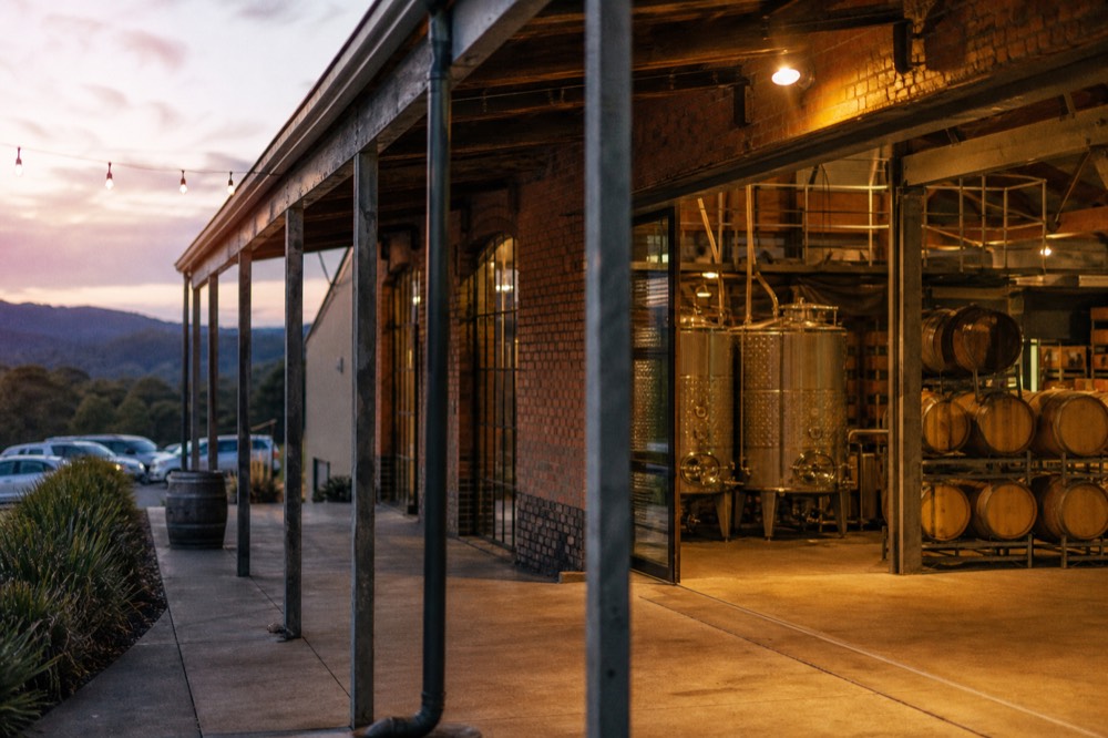Stainless steel wine tanks and stacked oak barrels inside a converted heritage warehouse with exposed brick and industrial windows