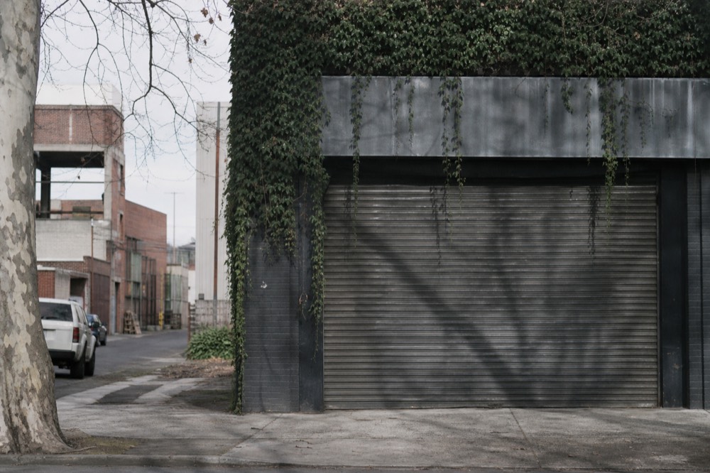 Empty shuttered warehouse frontage in an inner northern Melbourne street, roller door down, plane tree shadow across the concrete