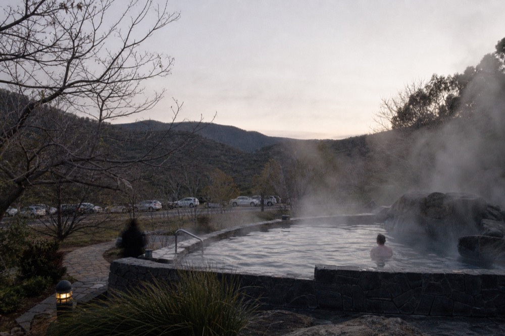 Steam rising from an outdoor thermal bathing pool set into a hillside at dusk with bare winter trees around it