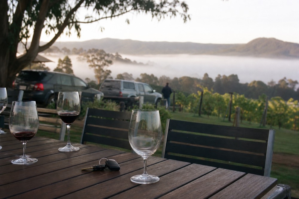 Car keys and a single untouched wine glass on an outdoor table beside a vineyard, other glasses filled nearby