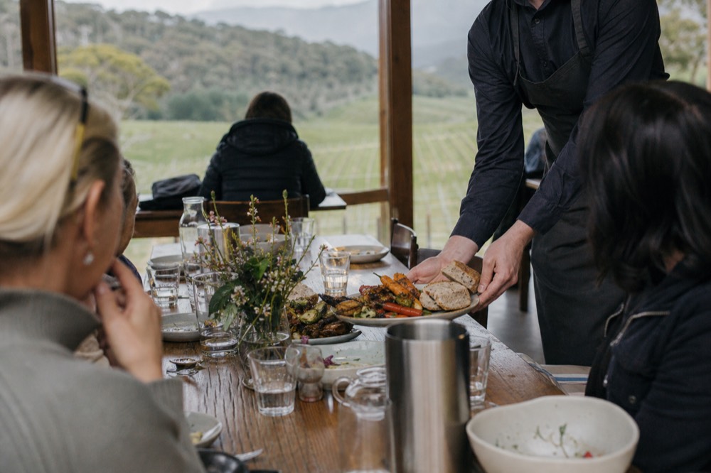 Waiter placing a share plate of roasted vegetables and bread on a long table of tour guests inside a vineyard restaurant